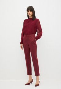 Blusa plissettata bordeaux con maniche semi-trasparenti abbinata a pantaloni bordeaux su misura e décolleté a punta coordinati con accenti dorati.