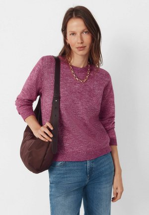 Fioletowy dziergany sweter z prążkowanym wykończeniem na dole i mankietach, zestawiony z ciemnobrązową torebką na ramię. Modelka nosi złoty łańcuszek; widoczne są dżinsy.