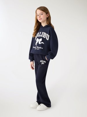 Młoda dziewczyna ubrana w granatową bluzę z kapturem "Malibu Santa Monica" oraz pasujące spodnie dresowe, stoi na jednolitym białym tle.