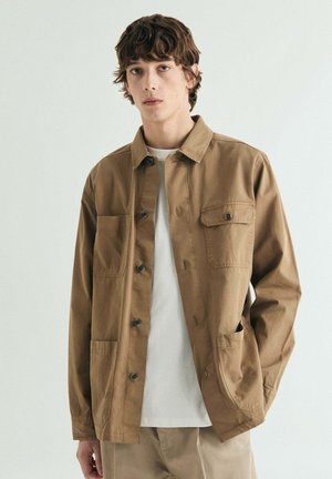 Joven con cabello rizado y castaño que lleva una chaqueta marrón claro abotonada sobre una camisa blanca, de pie frente a un fondo claro y liso.