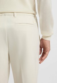 Licht beige broek met een gladde textuur, voorzien van een achterzak en een subtiele logo-accent. De stof lijkt ademend en op maat gemaakt.