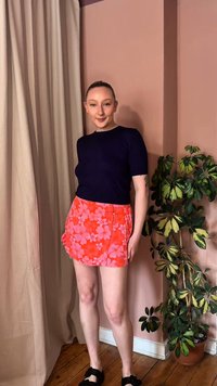 Γυναίκα στέκεται σε εσωτερικό χώρο με μαύρο κοντομάνικο τοπ, κόκκινη μίνι φούστα με φλοράλ σχέδιο και μαύρα παπούτσια δίπλα σε γλαστρωτό φυτό με ποικιλόχρωμα φύλλα.