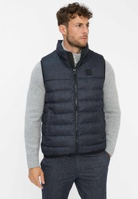 Dunkelblau gesteppte Weste mit hohem Kragen, Reißverschluss und einem kleinen Logo-Patch. Getragen über einem hellgrauen Pullover und gemusterten Hosen.