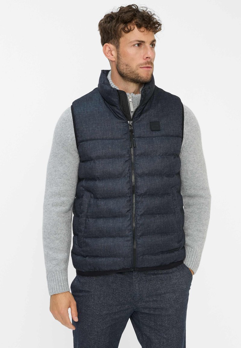 Dunkelblau gesteppte Weste mit hohem Kragen, Reißverschluss und einem kleinen Logo-Patch. Getragen über einem hellgrauen Pullover und gemusterten Hosen.