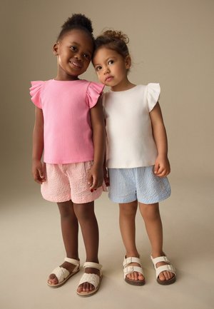 Deux jeunes filles debout côte à côte, l'une portant un haut à manches volantées rose et un short rayé, l'autre un haut à manches volantées blanc et un short bleu.