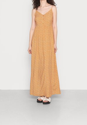 Mujer que lleva un vestido maxi sin mangas naranja con un pequeño estampado floral blanco y sandalias blancas con suelas negras gruesas.