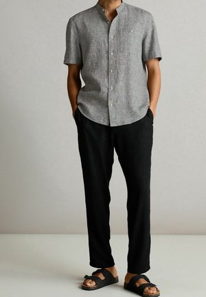 Homme portant une chemise grise à manches courtes boutonnée, un pantalon noir et des sandales noires à deux brides, debout les mains dans les poches.