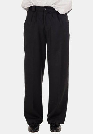 Pantaloni neri a vita alta con gamba larga e passanti per cintura indossati con mocassini neri con nappine su sfondo bianco.