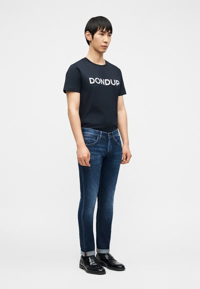Νεαρός άνδρας που φοράει μαύρο t-shirt με το λογότυπο Dondup, σκουρό μπλε στενά τζιν με γυρισμένα μπατζάκια και μαύρα δερμάτινα loafers, στέκεται μπροστά σε απλό φόντο.