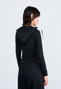 Sweatshirt preto com riscas brancas laterais, design ajustado, mangas longas e cordão ajustável. Textura suave e detalhes mínimos.