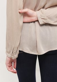 Blouse beige à manches longues en tissu doux et texturé, avec un ourlet arrondi et des poignets à boutons, présentée aux côtés d'un jean foncé.