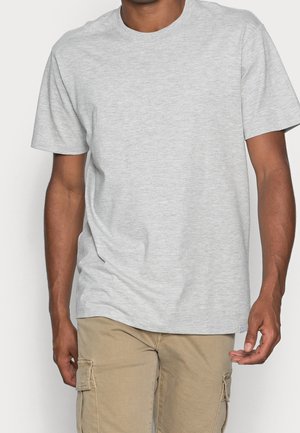 Mężczyzna w prostym jasnoszarym t-shircie z krótkim rękawem i beżowych spodniach cargo, stojący na neutralnym tle.