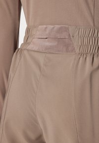 Hnědé elastické kalhoty z látky s hladkým povrchem, se širokým elastickým páskem a síťkovou kapsou na zadní straně.