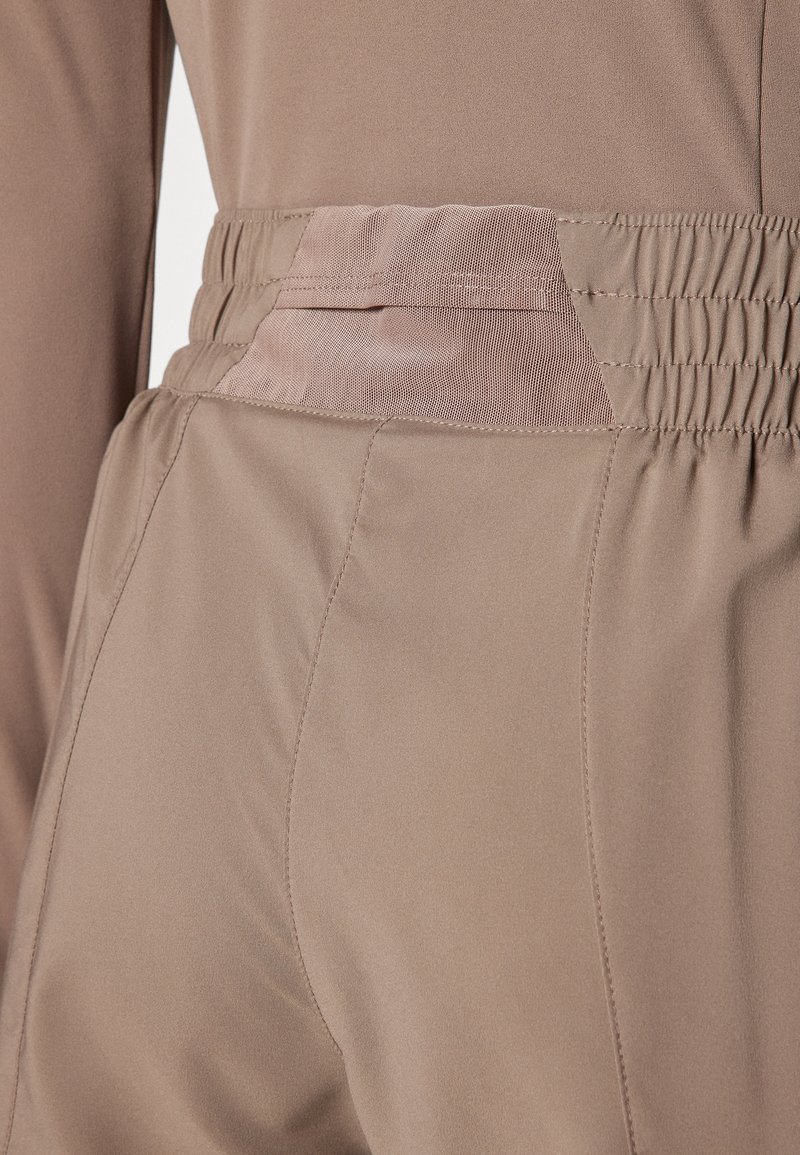 Pantaloni in tessuto elasticizzato marroni con una texture liscia, dotati di un elastico in vita e di un dettaglio in rete sulla tasca posteriore.