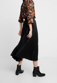 Svart plisserad kjol med en flödande design, kombinerad med en genomskinlig blus med blommönster i livfulla färger och svarta ankelstövlar med klack.