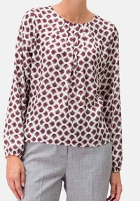 Femme portant un blouse blanche à manches longues avec un motif géométrique violet et des plis au niveau du col, associée à un pantalon gris clair.