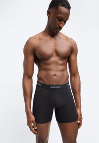 Skjorteløs mann med muskuløs kroppsbygning som ser ned, iført svarte Calvin Klein boxer shorts mot en enkel, lys bakgrunn.