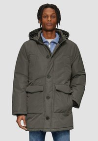 Donkergroene gevoerde parka-jack met een capuchon, vier voorzakken en een knoopsluiting, gedragen over een lichtblauw zip-up shirt.
