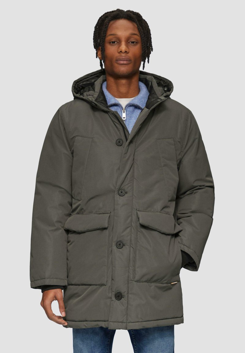 Donkergroene gevoerde parka-jack met een capuchon, vier voorzakken en een knoopsluiting, gedragen over een lichtblauw zip-up shirt.
