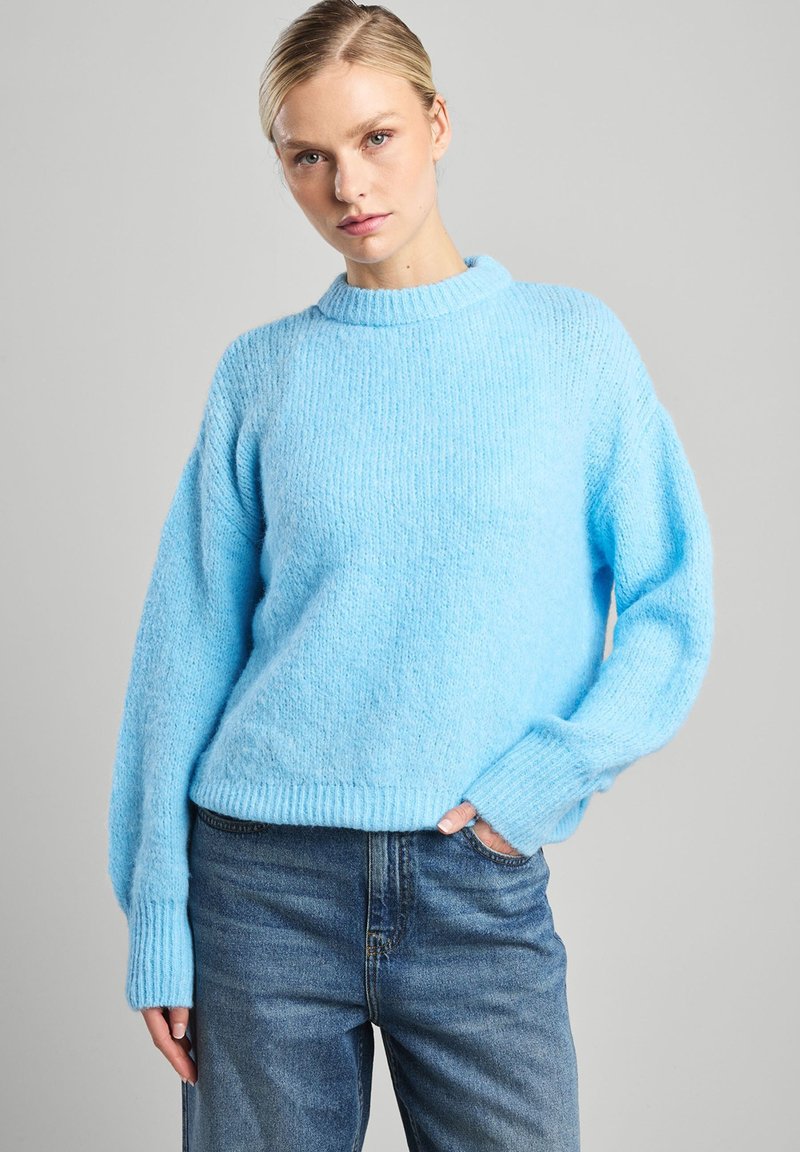 Pull en maille bleu clair avec un col et des poignets côtelés, offrant une coupe décontractée et une texture duveteuse, associé à un jean en denim.