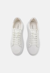 White sneakers with a smooth synthetic upper, round toe, flat rubber sole, and central lacing system. "Calvin" logo printed inside.