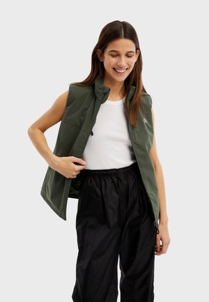 Chaleco verde sin mangas con cuello alto, que presenta una textura suave y un logotipo sutil, llevado sobre una camiseta blanca y pantalones negros.