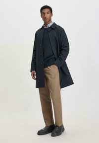 Marineblauer Trenchcoat mit Knopfverschluss, getragen über einem dunkelblauen Pullover und einem hellblauen Hemd. Beige Hosen und schwarze Stiefel runden das Outfit ab.