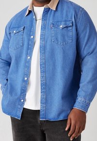 Camicia di denim blu con colletto in corduroy beige, due tasche sul petto e bottoni sulla parte anteriore. Indossata sopra una T-shirt bianca.