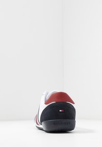 Vit sneaker med svart sula, svart häl och röd krage, som visar en liten röd, vit och blå Tommy Hilfiger-logotyp på den bakre hälen.