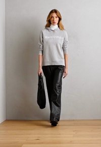 Grauer Kapuzenpullover mit weißem Logo, schwarze Kunstlederhosen und eine strukturierte schwarze Handtasche. Das Model steht auf einem Holzboden vor einer hellen Wand.