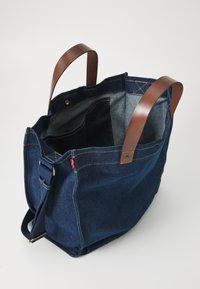 Denim draagtas met bruine leren handvatten. Interieur met grijze voering en meerdere zakken. Blauwe stof met zichtbare stikdetails.