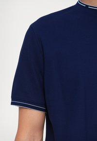 Tumedat sinine polo särk, millel on struktureeritud kangas, lühikesed varrukad ja kaelus, millel on kontrastsed valged triibud. Nähtav õlavarre õmbluse detail.