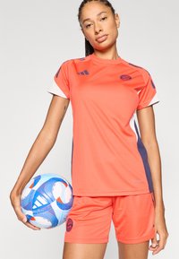 Φωτεινή πορτοκαλί αθλητική φανέλα με λευκές και ναυτικές λεπτομέρειες, σετ με αντίστοιχα σορτς. Παίκτης κρατά μια μπλε και λευκή μπάλα ποδοσφαίρου με γραφικά σχέδια.