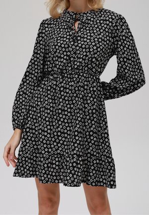 Kobieta ubrana w czarną sukienkę z długim rękawem, ozdobioną białymi wzorami gwiazd, z dekoltem typu keyhole, stojąca na jednolitym tle.