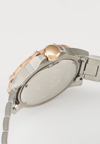 Montre en métal argenté et or rose avec une lunette texturée, une couronne proéminente et un bracelet en métal. L'arrière présente des marquages gravés et une finition élégante.