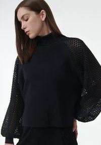 Zwarte sweatshirt met een hoge hals, een solide lichaam en volumineuze, textuurvolle mesh mouwen. De stof is zacht met een modern ontwerp.