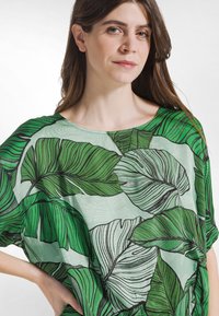 Transparent, let top med et mønster af grønne blade på en lys baggrund; løs, oversized pasform; bred halsudskæring og korte ærmer.