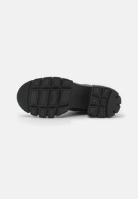 Suela de zapato deportivo negro con patrones de dibujo profundos, material de goma, surcos rectangulares y circulares para tracción, bordes ligeramente elevados.