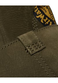 Detalle de bota de cuero verde oliva con una correa cosida y acabado texturizado, con una etiqueta de tela azul y amarilla parcialmente visible.