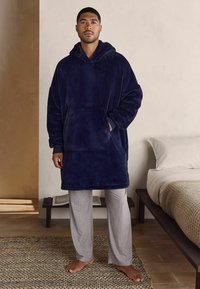 Námornícky modrý, oversized fleece mikina s kapucňou a prednou vreckom, nosená cez svetlo šedé, pruhované tepláky. Mäkký povrch.