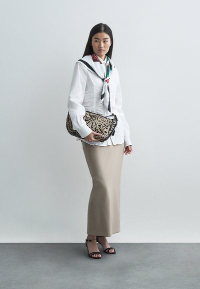 Kobieta w białej bluzce z marszczeniami, beżowej spódnicy, kolorowej apaszce, czarnych sandałach, trzymająca kopertówkę w panterkę na tle szarego tła.