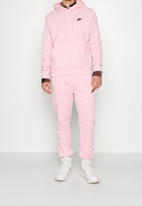 Rosa Nike-hoodie och matchande sweatpants i mjukt fleecematerial, med en svart logga på bröstet och sidan. Bärs med vita sneakers.