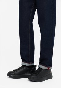 Sneakers neri con lacci, in pelle liscia, punta tondeggiante e suola testurizzata, abbinati a jeans in denim scuro con orli grigi risvoltati.