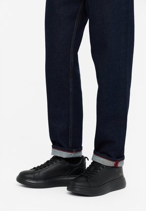 Sneakers neri con lacci, in pelle liscia, punta tondeggiante e suola testurizzata, abbinati a jeans in denim scuro con orli grigi risvoltati.