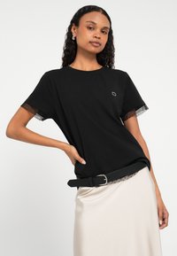 Črna majica s kratkimi rokavi, okrašena z izrezom iz prozorne mrežaste tkanine. Majica ima okrogel izrez in majhen zlati logotip na prsih.