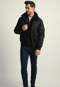 Mężczyzna w czarnej kurtce bombowcu z kołnierzem z owczej wełny, założonej na czarny sweter z zamkiem błyskawicznym i ciemnogranatowych spodniach, stoi z ręką na suwak kurtki.