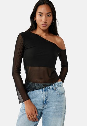 Mujer con top negro transparente de un solo hombro y manga larga y vaqueros azul claro de talle alto, de pie frente a un fondo liso.
