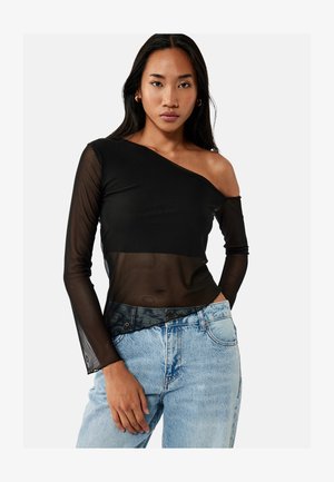 Femme portant un haut transparent noir à une épaule avec manches longues et un jean taille haute bleu clair, debout devant un fond uni.