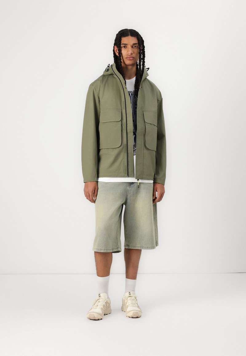 Chaqueta verde oliva con capucha, con dos grandes bolsillos delanteros, combinada con pantalones cortos de mezclilla claros y zapatillas beige. Diseño simple; ajuste informal.