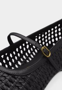 Μαύρο πλεκτό επίπεδο παπούτσι με διάτρητες πλευρές και λεπτό λουράκι που στερεώνεται με μια μικρή, γυαλιστερή χρυσή αγκράφα στο πλάι.